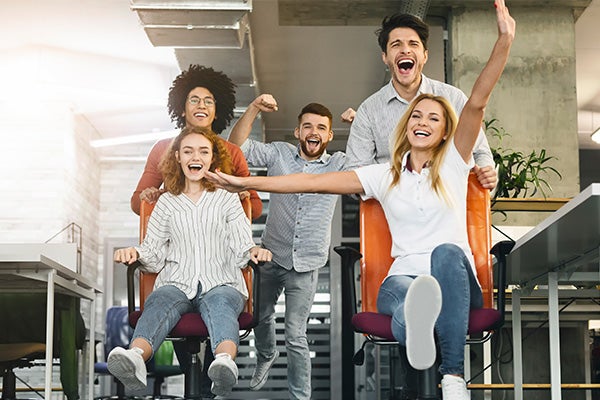 Group of cheerful young coworkers racing in office chairs and laughing in a modern workspace.