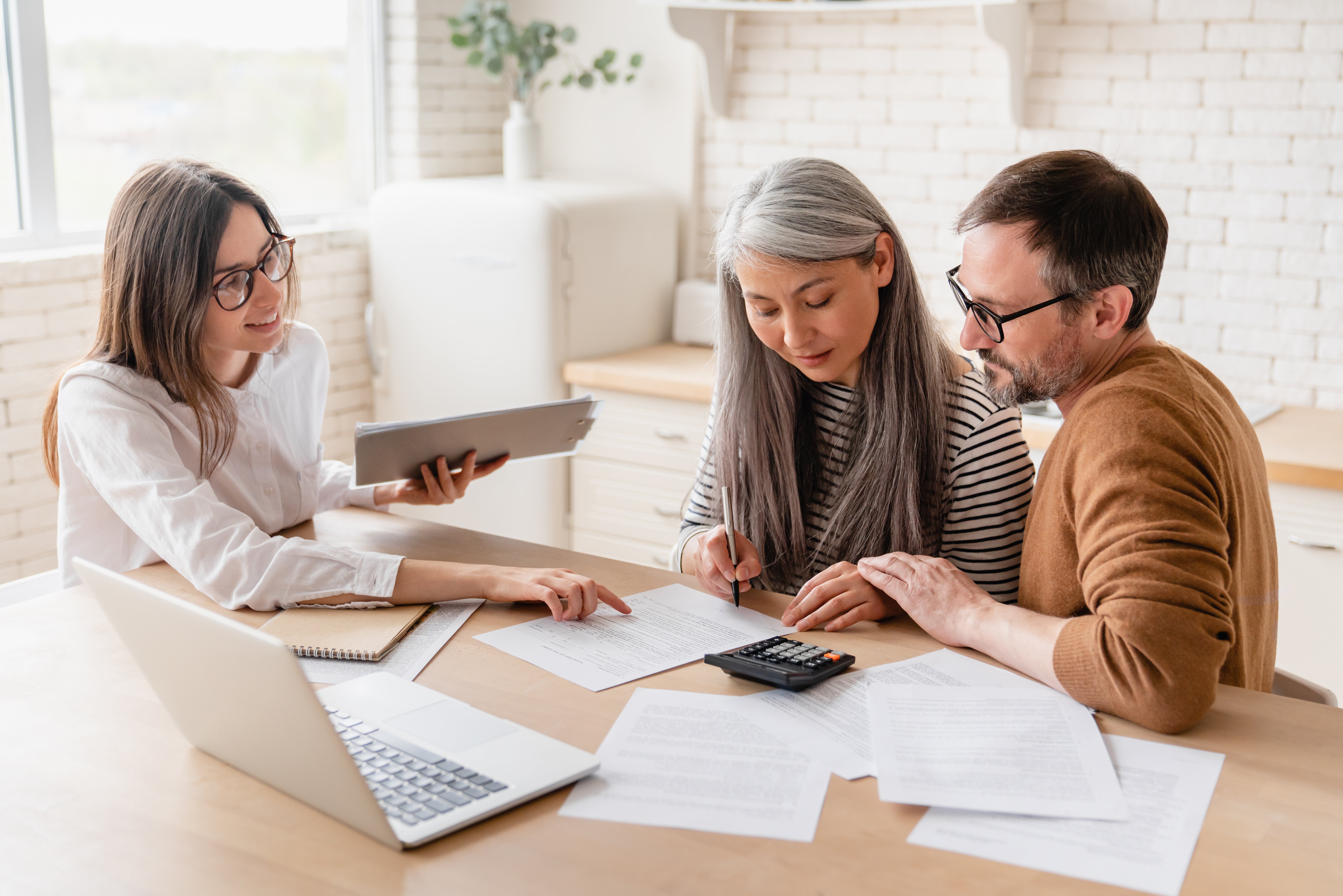 A couple reviews documents and uses a calculator while meeting with a financial advisor at a table with a laptop.
