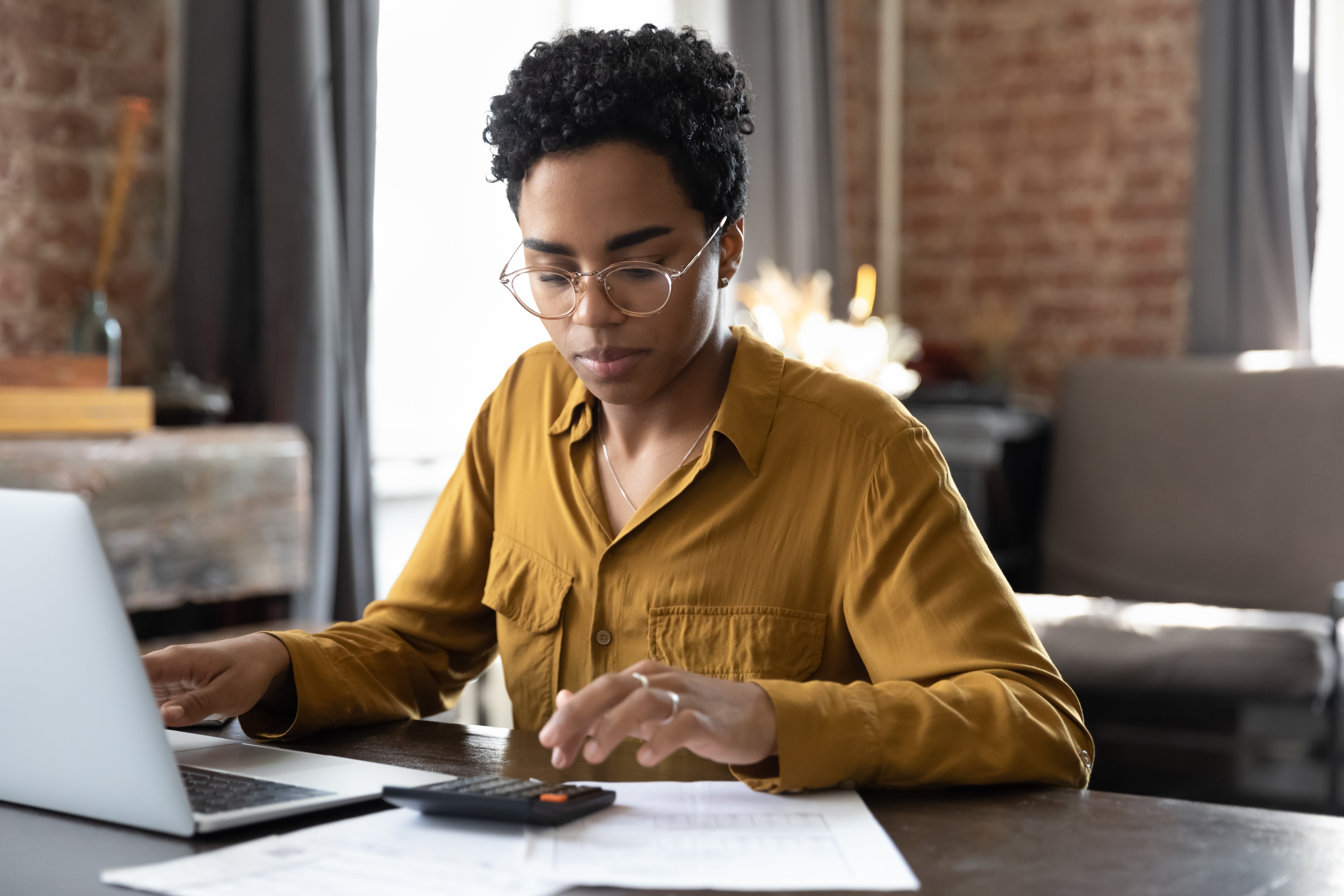 Person wearing glasses and a mustard shirt using a calculator while working on a laptop at a desk.