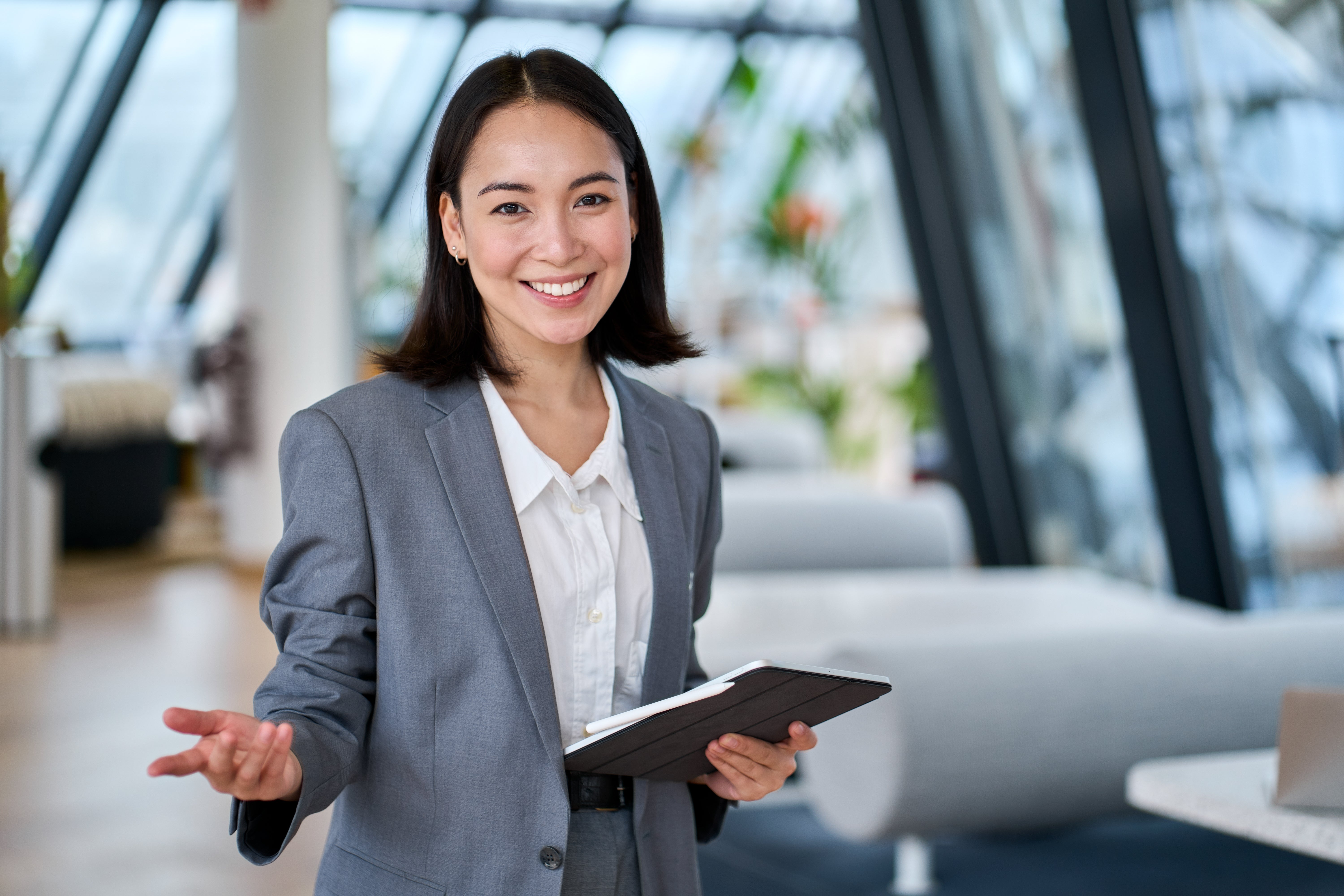 A smiling professional woman in a gray suit gestures while holding a digital tablet in a modern office setting.