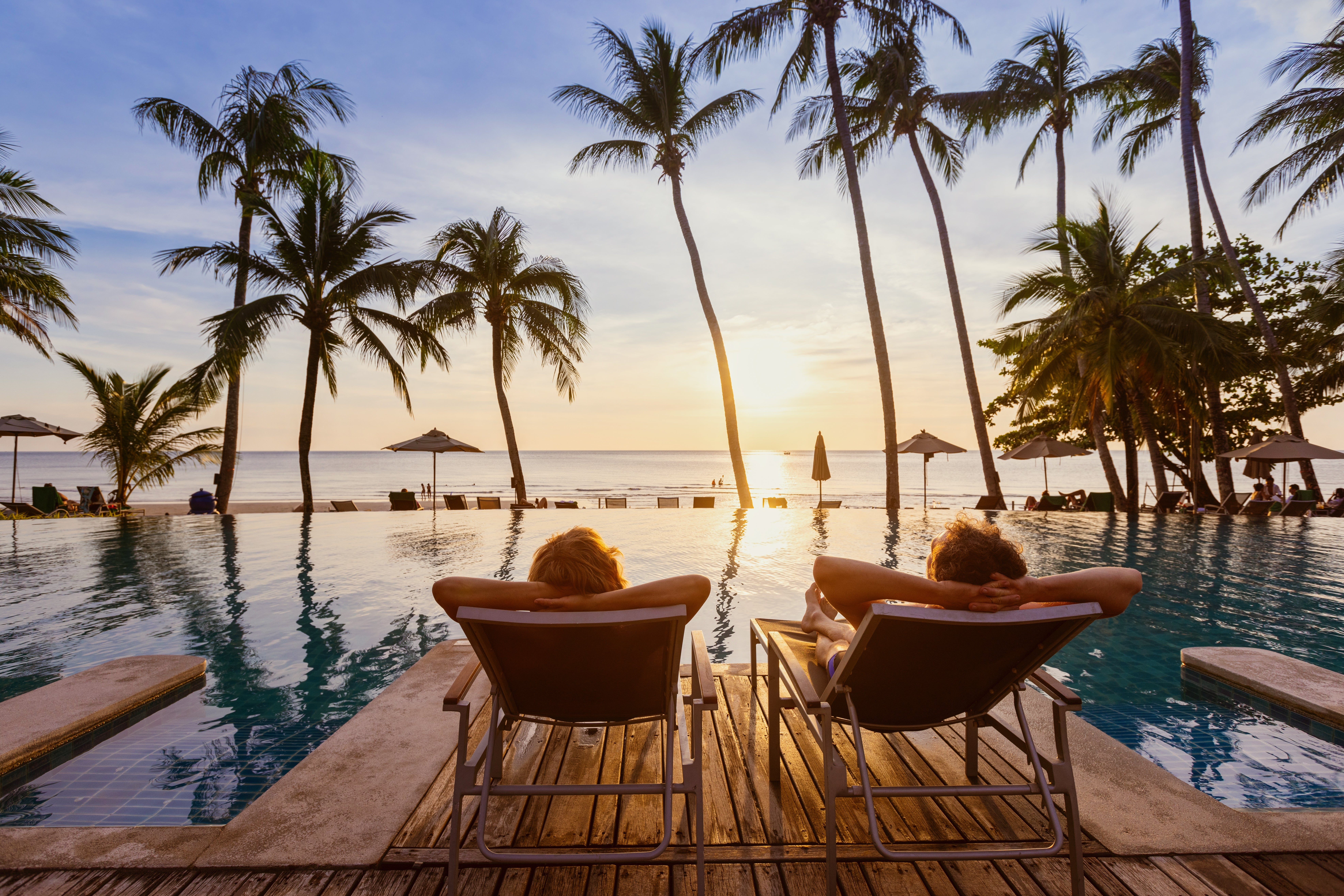 Two people relaxing on lounge chairs by a tropical pool, facing the ocean as the sun sets behind palm trees.