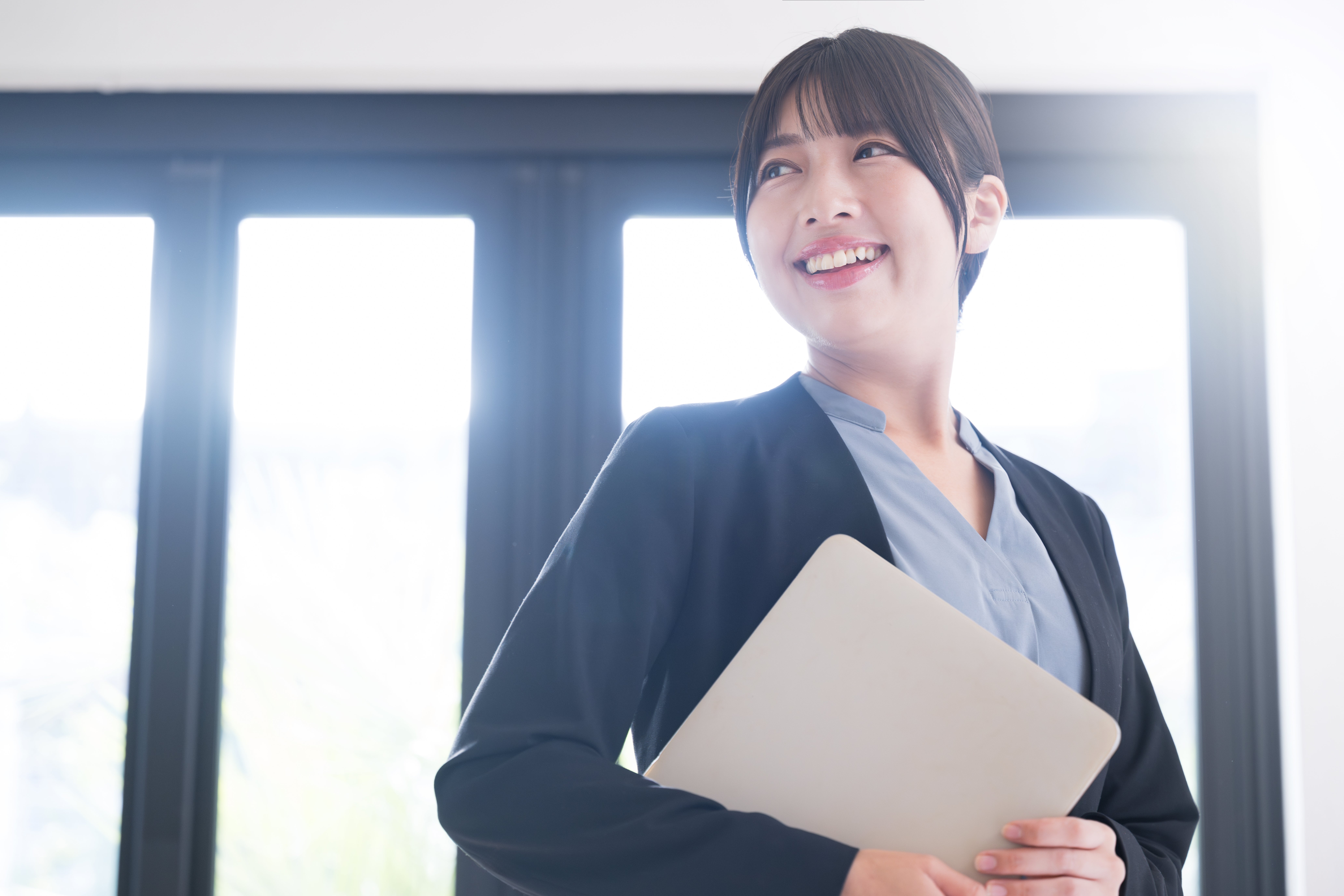 A professional woman smiling confidently while holding a closed laptop, standing in front of large windows with natural light streaming in.